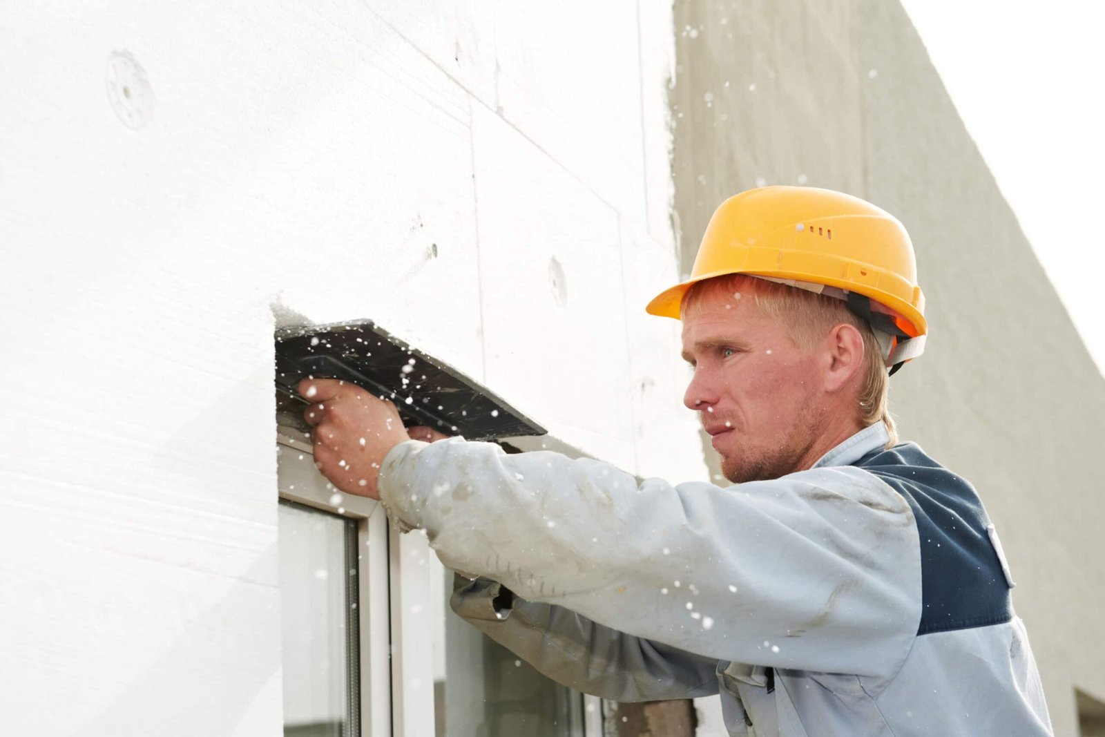 Un ouvrier du bâtiment portant un casque de sécurité jaune utilise habilement une truelle pour lisser le matériau isolant blanc sur un mur extérieur. Des particules de neige ou de mousse flottent dans lair tandis que le façadier concentré, vêtu dune veste bleu clair, effectue sa tâche méticuleuse.